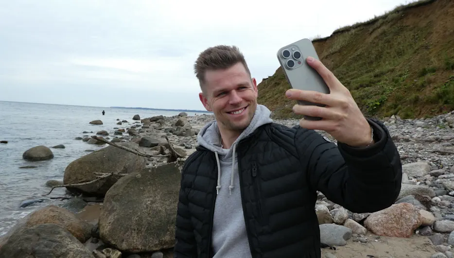 Ein Mann in schwarzer Jacke macht ein Selfie mit einem grauen Smartphone am steinigen Strand. Im Hintergrund sind Meer und Klippen.