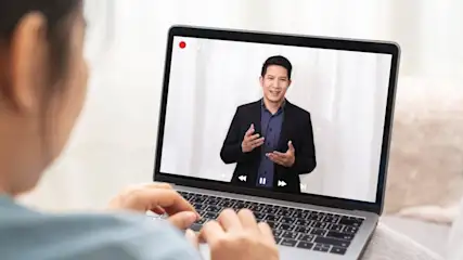 Eine Person tippt auf einem Laptop, auf dessen Bildschirm ein Mann in einem schwarzen Sakko gestreamt wird.