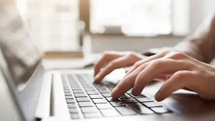 Hände tippen auf einer Laptop-Tastatur vor einem hellen, unscharfen Hintergrund.