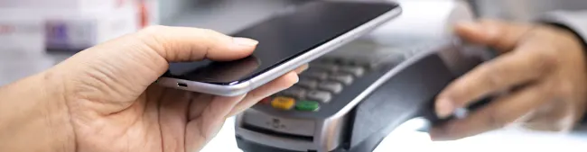 Eine Hand hält ein schwarzes Smartphone über ein graues Zahlungsgerät mit Tasten in Gelb und Grün.