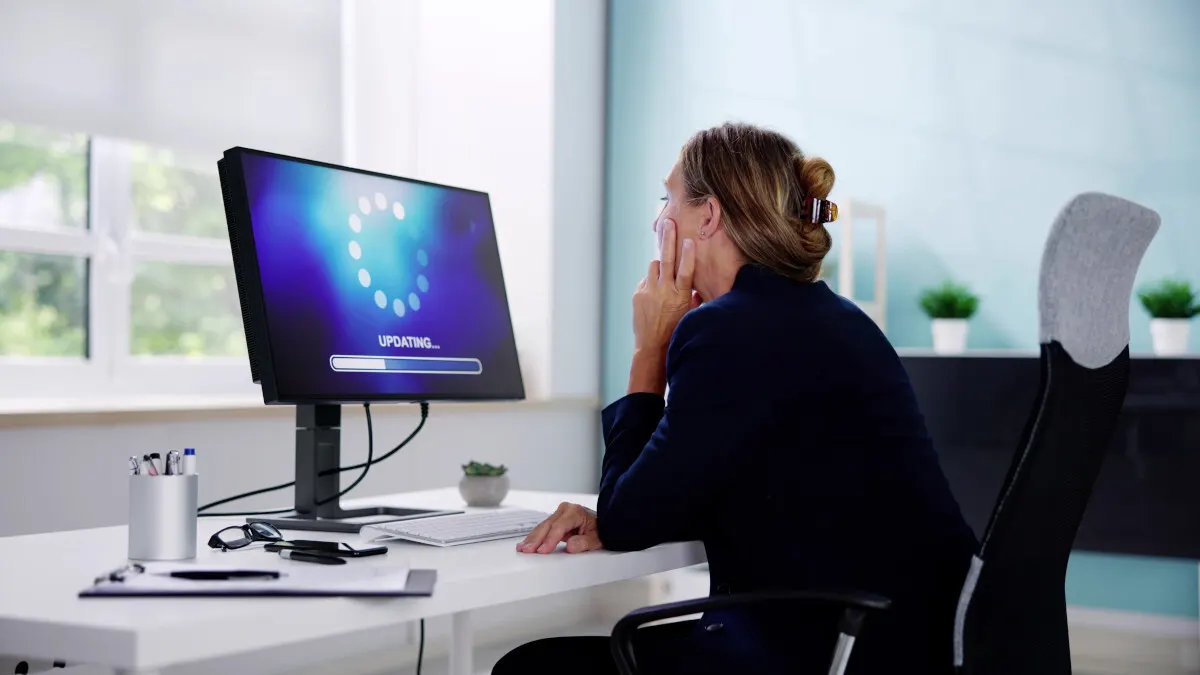 Eine Frau sitzt vor einem Computer, der ein Update anzeigt, mit Büromaterialien auf einem weißen Schreibtisch.