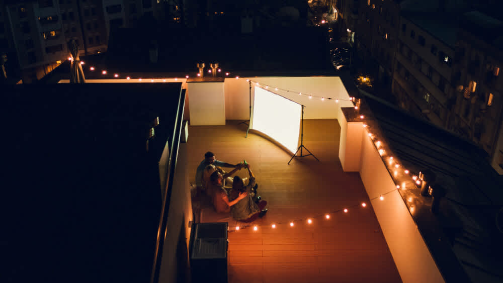 Eine Gruppe von Menschen sitzt abends auf einer Dachterrasse und schaut auf eine erleuchtete Beamer-Leinwand.
