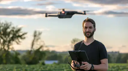 Ein Mann hält eine Steuerung für eine Drohne in einem Feld mit Bäumen und einem bewölkten Himmel im Hintergrund.