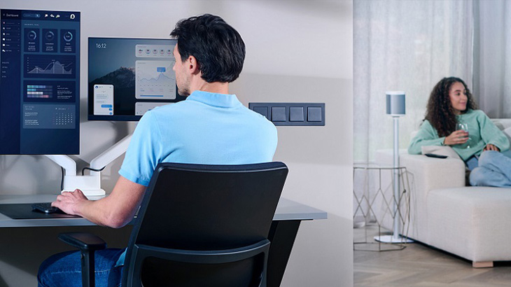 Ein Mann arbeitet an zwei Monitoren, während eine Frau auf einem Sofa sitzt und ein Glas hält.