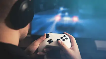 Ein Mann mit Kopfhörern hält einen weißen Controller vor einem Bildschirm mit blauer Grafik und roten Lichtern.