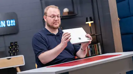 Ein Mann mit Brille hält ein silbernes Tablet. Ein Tisch mit roter Auflage steht davor, im Hintergrund sind Regale und eine Lampe.