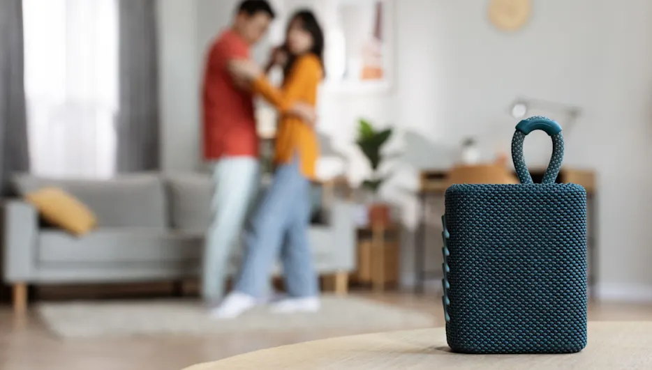 Ein blauer Lautsprecher steht auf einem Tisch. Im Hintergrund tanzt ein Paar in einem Wohnzimmer mit Sofa und Pflanze.