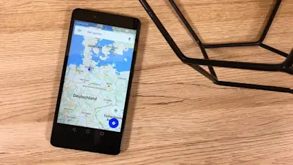 Ein schwarzes Smartphone zeigt eine Kartenanwendung mit Deutschland und Dänemark auf einem Holztisch neben einem schwarzen Metallobjekt.