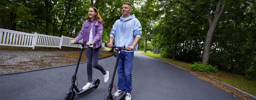Zwei Personen fahren nebeneinander auf E-Scootern über einen asphaltierten Weg im Grünen.