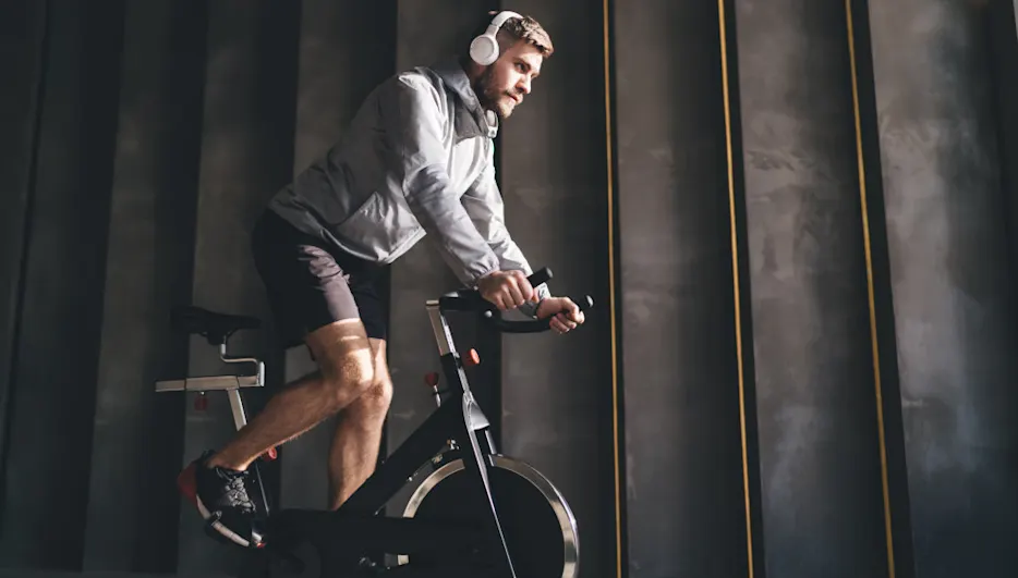 Ein Mann mit Kopfhörern fährt auf einem schwarzen Standfahrrad vor einer grauen Wand mit goldenen Streifen.