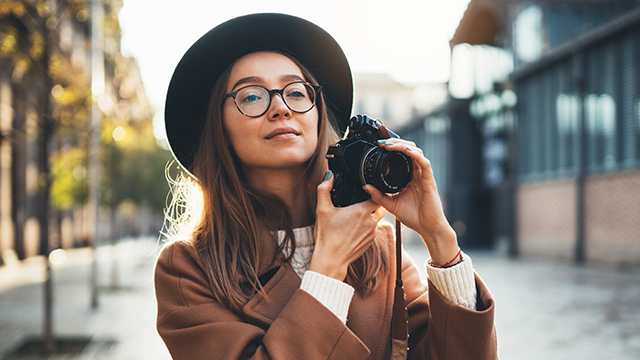 Eine Frau mit Hut und Brille hält eine schwarze Kamera in einer städtischen Umgebung. Bäume und Gebäude sind im Hintergrund sichtbar.