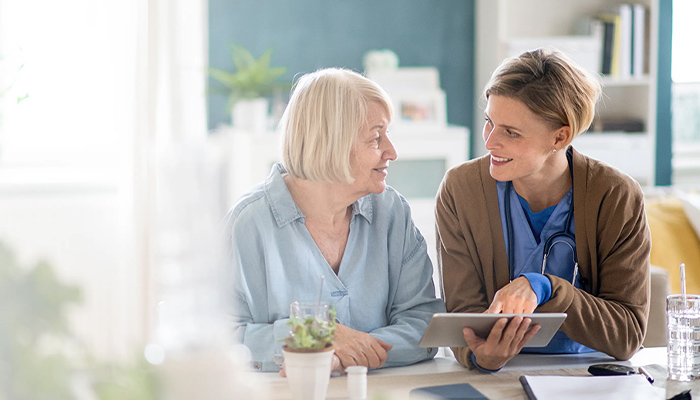 Pflegeperson mit Stethoskop zeigt einer älteren Frau ein Tablet; wohnliche Umgebung; Tisch mit Glas, Unterlagen und Pflanze. 