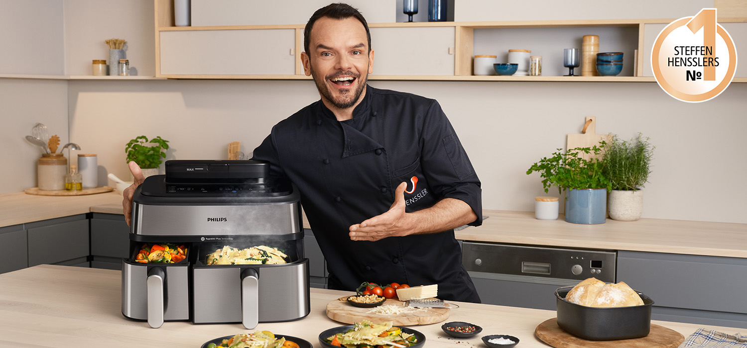Koch Steffen Henssler steht neben einem Philips Airfryer mit zwei Körben in einer Küche. Verschiedene Speisen sind vor ihm aufgebaut. Ein Logo mit der Aufschrift "Steffen Hensslers No. 1" ist abgebildet.