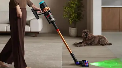 Eine Person saugt ein Wohnzimmer mit dem Dyson V16 Piston Animal, im Hintergrund liegt ein brauner Hund.