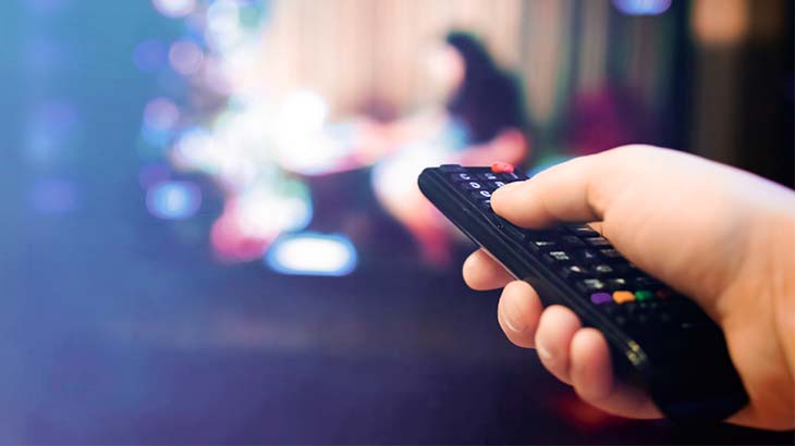 Eine Hand hält eine schwarze Fernbedienung vor einem verschwommenen Fernsehbildschirm mit blauen und roten Lichtern.