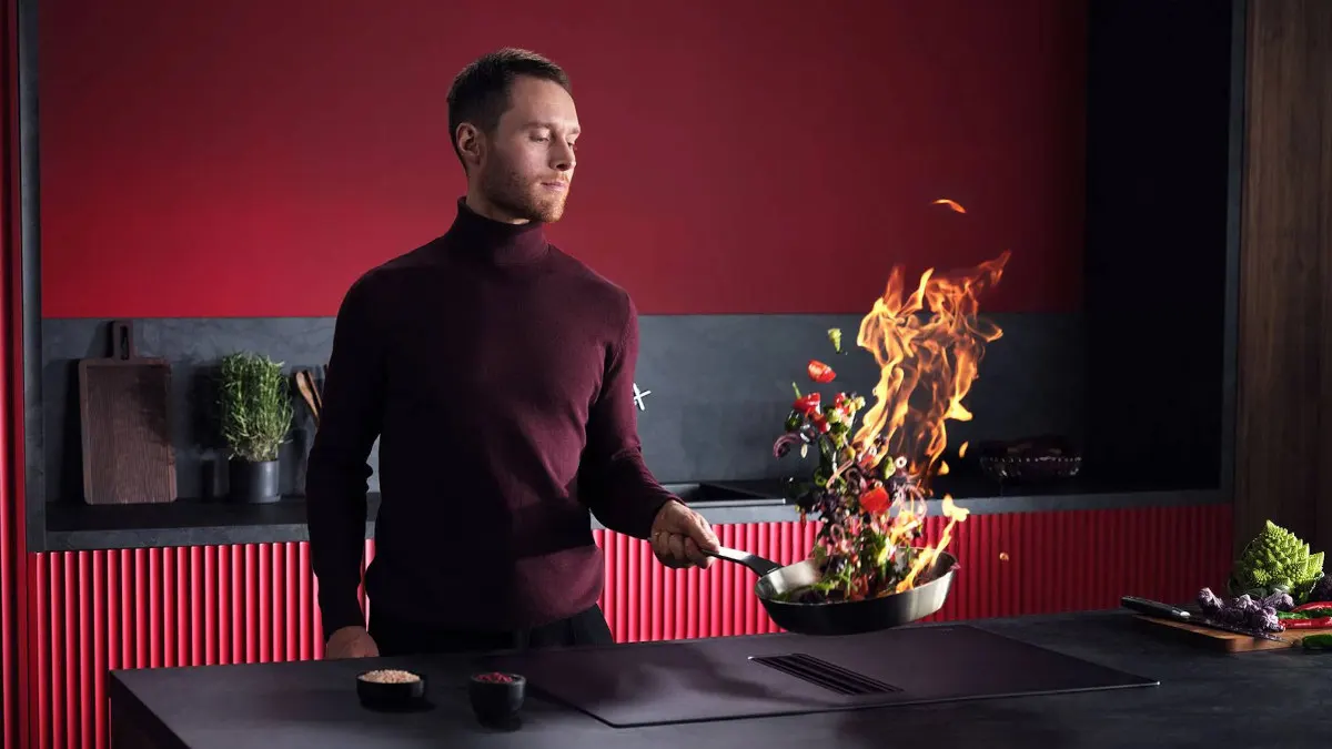 In einer Küche wirft ein Mann Gemüse in einer Bratpfanne in die Luft, Flammen steigen auf.