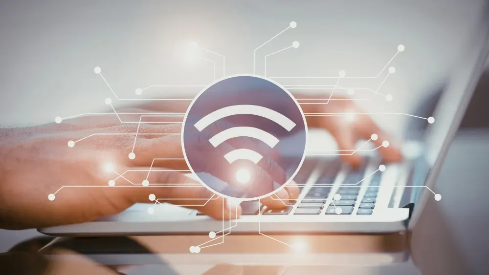 Eine Person tippt auf einem Laptop, überlagert von einem WLAN-Symbol und einem Netzwerk aus Linien und Punkten.