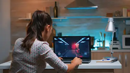 Eine Person spielt ein Videospiel auf einem Laptop in einer Küche mit Regalen und Geräten im Hintergrund.