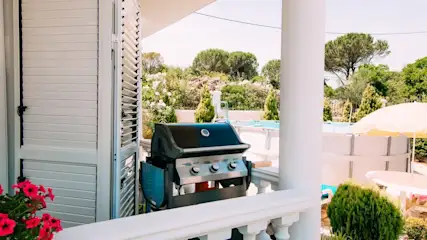 Außenansicht mit Grill auf Veranda, Blick auf Pool und Garten durch weiße Säulen und Geländer. Blühende Pflanzen im Vordergrund.
