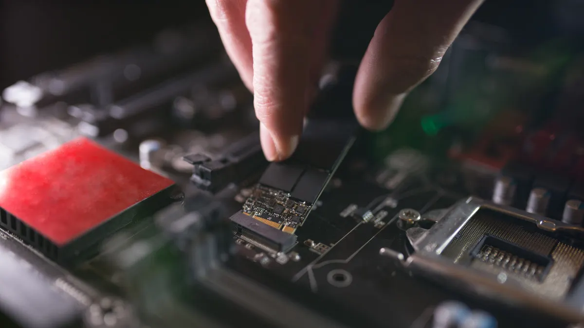 Eine Hand setzt eine schwarze SSD-Karte in eine Computer-Hauptplatine mit roten und silbernen Komponenten ein.