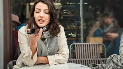 Eine Frau mit Kopfhörern hält ein Smartphone am Tisch in einem Café im Freien.