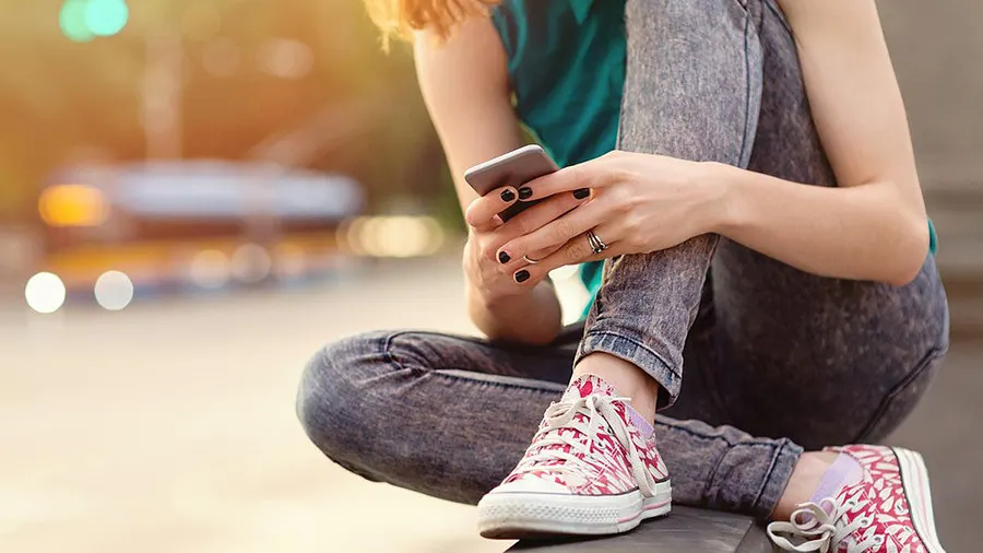 Eine Person sitzt mit gekreuzten Beinen und hält ein Smartphone in der Hand, Jeans und gemusterte Turnschuhe, verschwommener Hintergrund.
