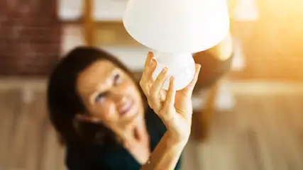 Eine Frau hält eine weiße Lampe in der Hand, im Hintergrund sind eine Wand und Regale zu sehen.