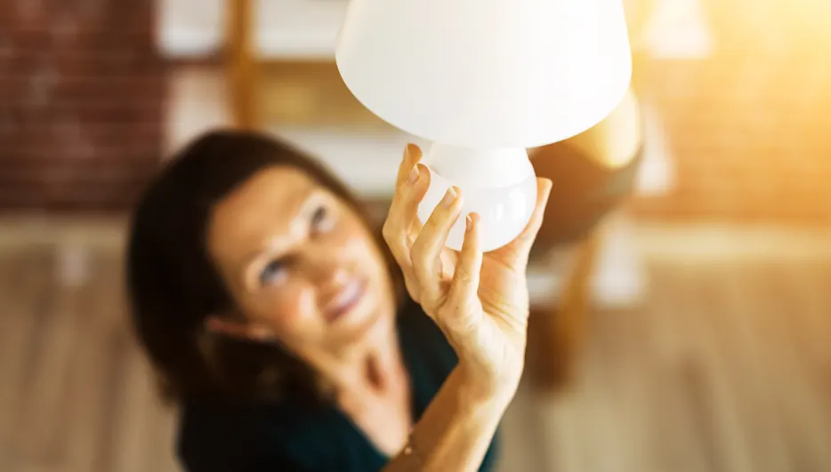 Eine Frau hält eine weiße Lampe in der Hand, im Hintergrund sind eine Wand und Regale zu sehen.