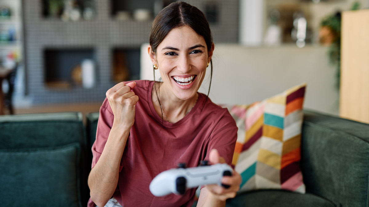 Eine Frau sitzt auf einer Couch. Sie hat einen Controller in der einen Hand und jubelt mit der anderen.