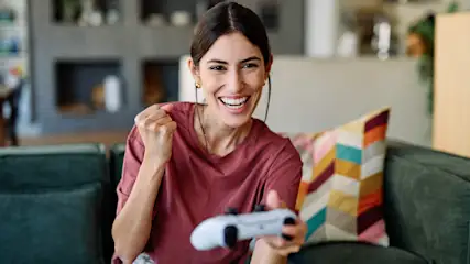 Eine Frau sitzt auf einer Couch. Sie hat einen Controller in der einen Hand und jubelt mit der anderen.