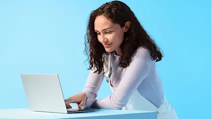 Eine Frau tippt auf einem silberfarbenen Laptop an einem türkisfarbenen Tisch vor einem hellblauen Hintergrund.