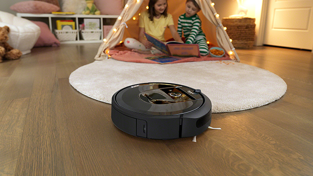 Ein schwarzer Staubsaugerroboter steht auf einem Holzboden vor einem runden, hellen Teppich. Zwei Kinder sitzen in einem Zelt.