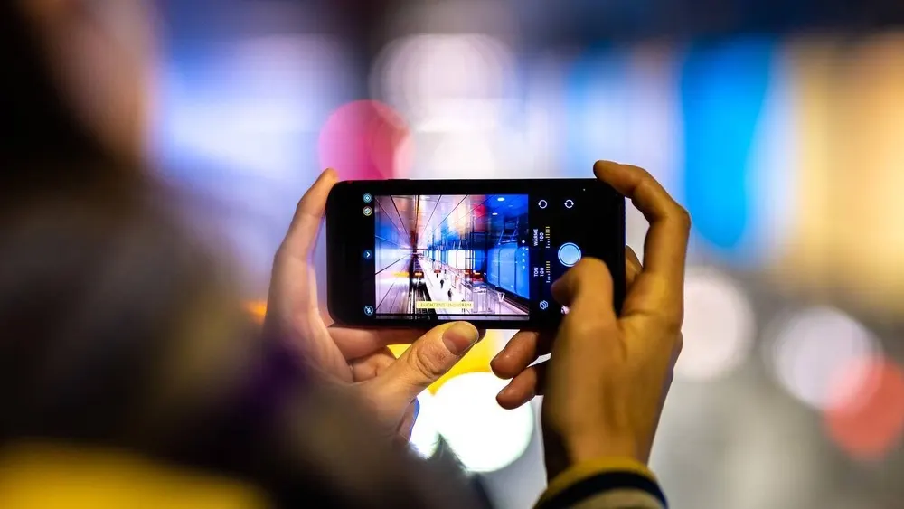 Eine Person hält ein Smartphone, um ein Foto von einem belebten Bahnhof mit blauem und rotem Licht zu machen.