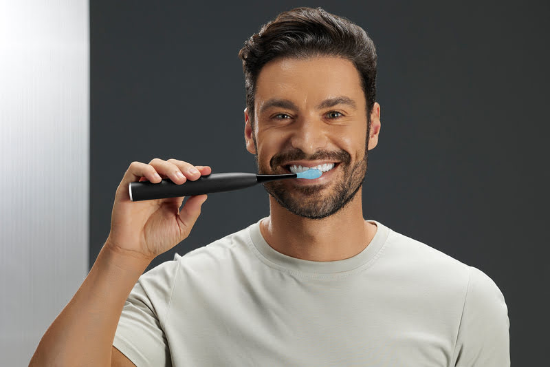 Ein Mann putzt sich die Zähne mit einer schwarzen elektrischen Zahnbürste vor einem grauen Hintergrund.