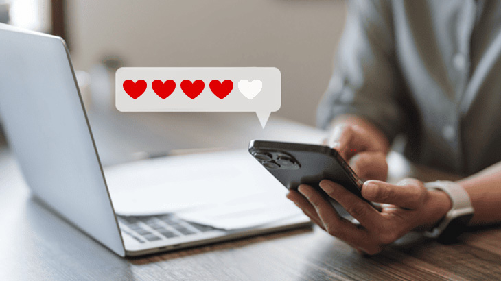 Person hält ein schwarzes Smartphone neben einem Laptop. Eine Sprechblase zeigt drei rote und ein leeres Herz.