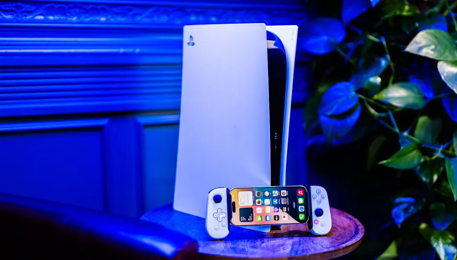 Ein weißes PlayStation 5-System steht auf einem Holztisch. Ein Smartphone mit Controllern liegt davor. Eine Pflanze ist im Hintergrund.