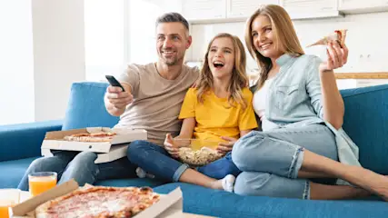 Eine Familie sitzt auf einem blauen Sofa, isst Pizza und Popcorn und schaut fern. Der Mann hält eine Fernbedienung.