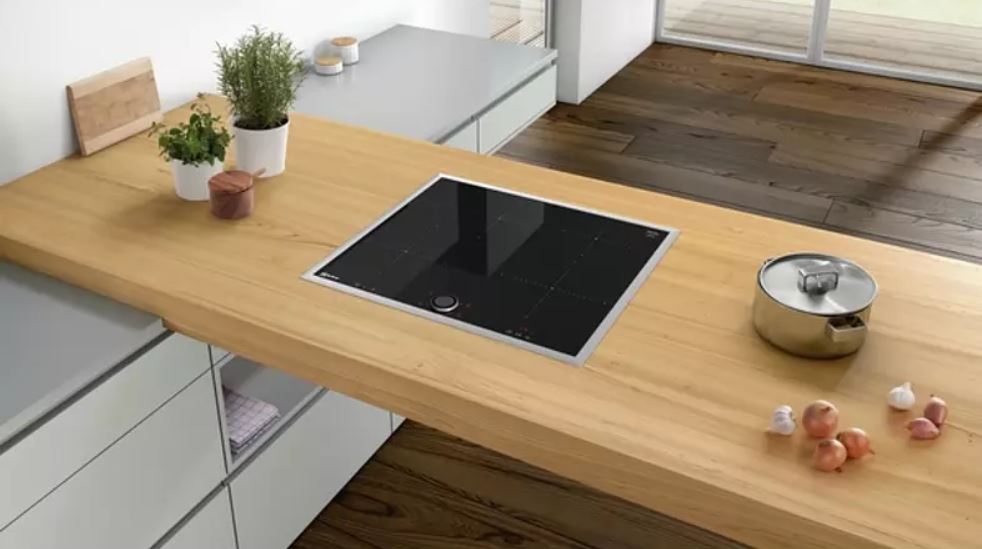 Auf einer Holzarbeitsplatte befinden sich ein schwarzes Kochfeld, ein Topf, Zwiebeln und Kräutertöpfe.