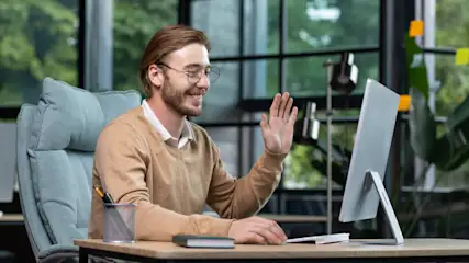 Ein Mann mit Brille winkt vor einem Computer an einem Schreibtisch mit Stifthalter und Notizbuch, vor einem Fenster.