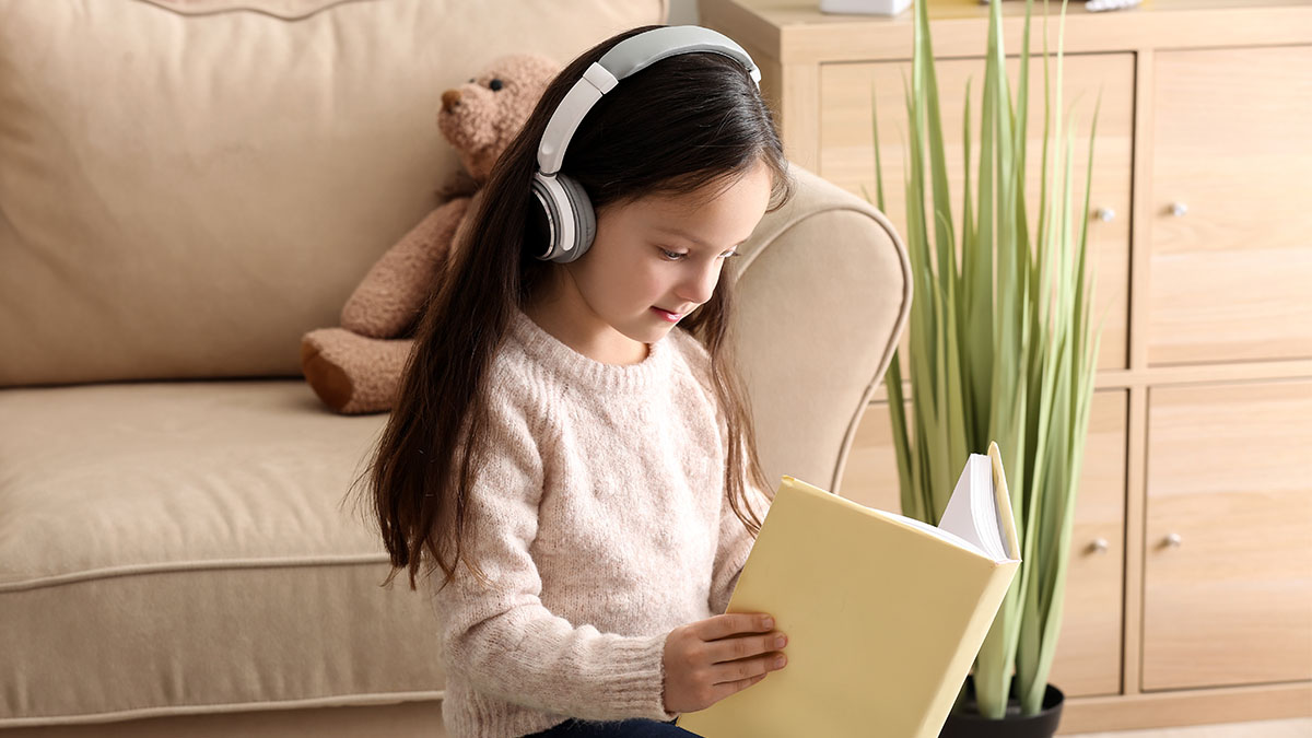 Ein Mädchen mit Kopfhörern sitzt vor einem Sofa und liest in einem gelben Buch. (Adobe Stock, Datei Nr. 249994490)
