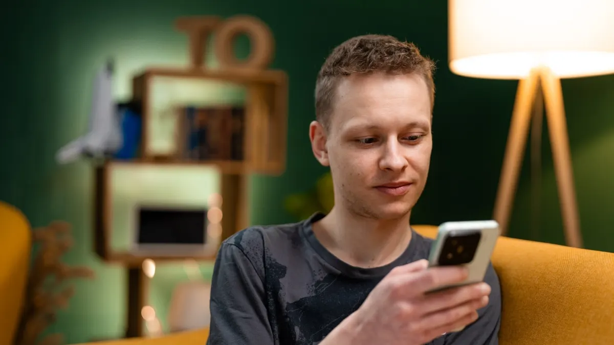 Ein Mann sitzt auf einem gelben Sofa und schaut auf ein weißes Smartphone in seiner Hand. Im Hintergrund sind ein Regal und eine Lampe.