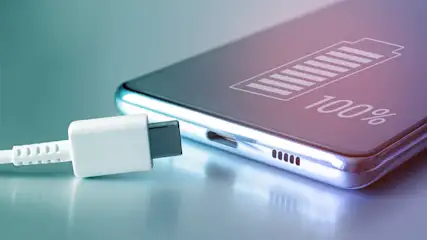 Ein Smartphone mit 100% Akku wird über ein weißes USB-C-Kabel aufgeladen. Das Display zeigt ein Batteriesymbol und "100%".