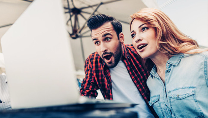 Ein Mann im karierten Hemd und eine Frau in Jeansjacke schauen überrascht auf einen Laptop.