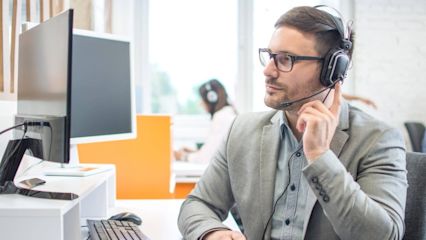 Mann sitzt im Büro und hält seinen Finger ans Headset
