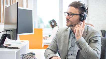 Mann sitzt im Büro und hält seinen Finger ans Headset