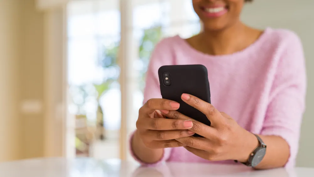 Frau im rosa Pullover hält ein schwarzes Smartphone in der Hand. Ein Armbanduhr ist am Handgelenk sichtbar.