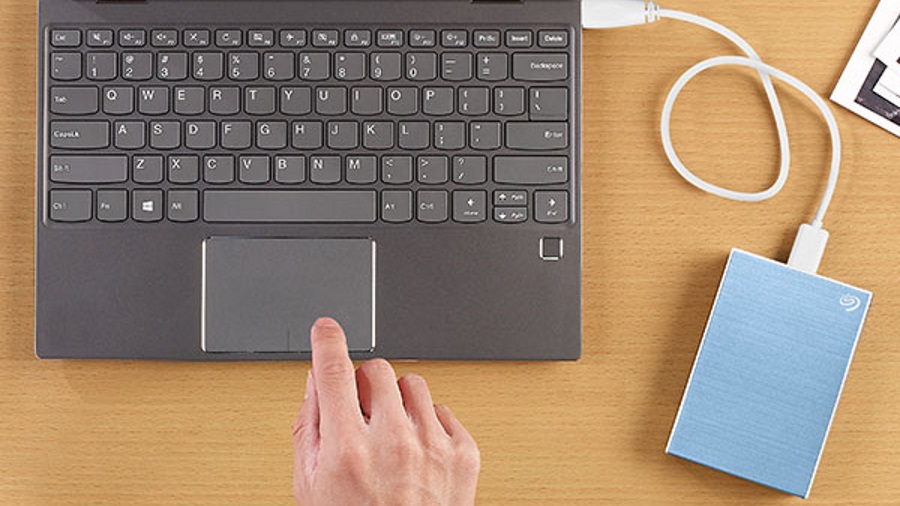 Ein grauer Laptop mit Tastatur und Touchpad, verbunden mit einer hellblauen externen Festplatte über ein weißes Kabel. Eine Hand berührt das Touchpad.