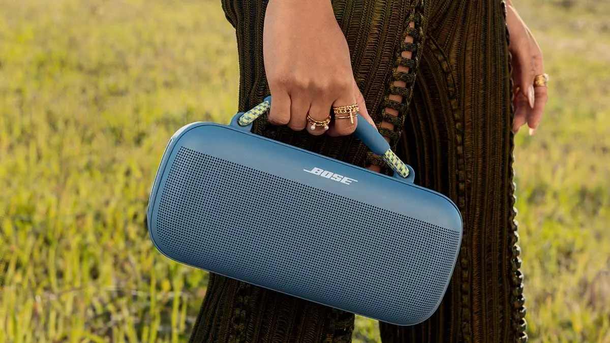 Eine Person hält einen blauen Bose-Lautsprecher mit einem Griff in der Hand, im Hintergrund ist Gras zu sehen.