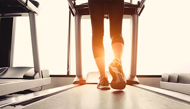 Beine einer Person in dunkler Hose und Sportschuhen auf einem Laufband vor einem hellen Fenster.