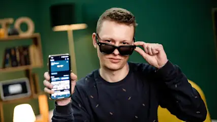 Mann mit Sonnenbrille hält ein Smartphone vor einem grünen Hintergrund mit Regalen und einer Lampe.
